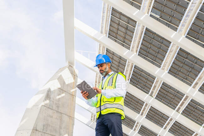 Niedriger Winkel eines hispanischen Ingenieurs mittleren Alters in Uniform mit Tablet, der auf den Bildschirm unter einem Solarkraftwerk blickt — Stockfoto