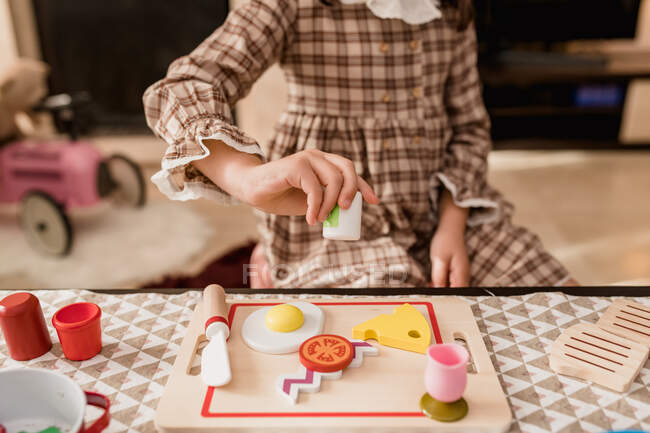 Recadrer enfant méconnaissable en robe à carreaux avec couteau jouet coupe fromage sur planche à découper tout en jouant dans la maison — Photo de stock