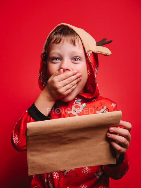 Shocked happy little boy festive costume covering opened mouth with hand and looking at camera while reading letter against red background during Christmas party — Stock Photo