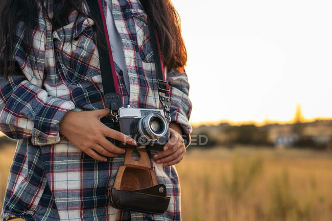 Crop donna irriconoscibile in camicia a scacchi che tiene vecchia macchina fotografica e custodia in pelle in retroilluminato — Foto stock