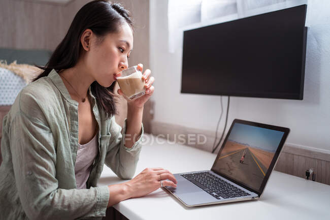 Young attentive ethnic woman enjoying coffee while playing video game on netbook at desk in house — Stock Photo
