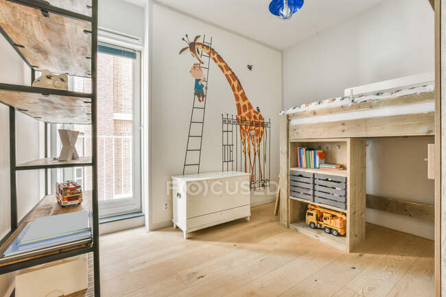 Cómodo interior de dormitorio para niños con foto de jirafa en la pared y estantes de madera con cama - foto de stock