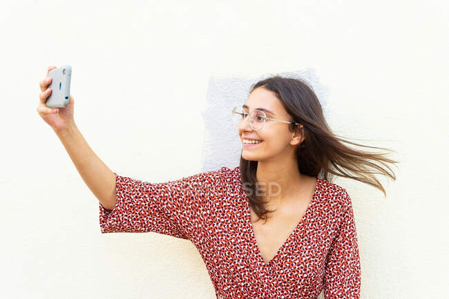 Улыбающаяся женщина в платье и очках стоит, делая селфи со смартфоном возле белой стены днем — стоковое фото