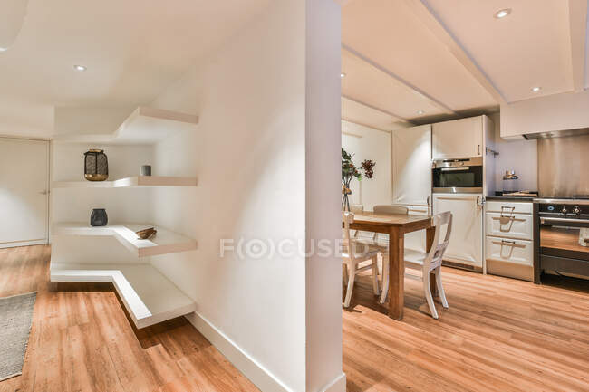 Интерьер современной кухни с обеденным столом и гостиной с полками, разделенными белой стеной в квартире — стоковое фото