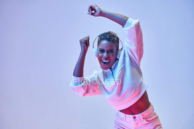 Енергетична етнічна жінка в бездротових навушниках і модний одяг танцює хіп-хоп з відкритим ротом дивлячись на камеру — стокове фото