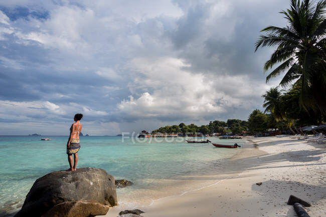 Полный вид босиком женщины-туристки в купальниках, стоящей на валуне и любующейся лазурным морем во время отдыха в Малайзии — стоковое фото