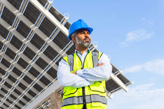 Low angle of ethnic male inspector in vest with protective helmet crossing arms standing against modern solar power station — Stock Photo