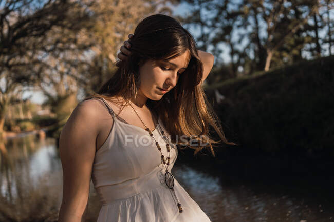 Suave adolescente en vestido de sol y cuentas tocando el cabello mientras mira hacia abajo contra el río en la luz del sol suave - foto de stock