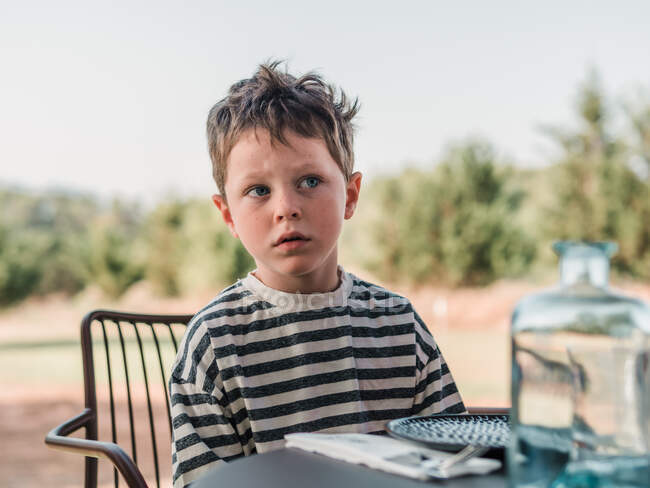 Очаровательный мирный ребенок сидит за столом во дворе летом и смотрит в сторону — стоковое фото