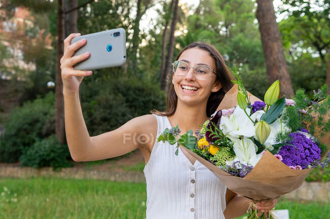Contenida joven hembra en gafas con flores florecientes ramo de flores tomando selfie en el teléfono celular en el parque urbano - foto de stock