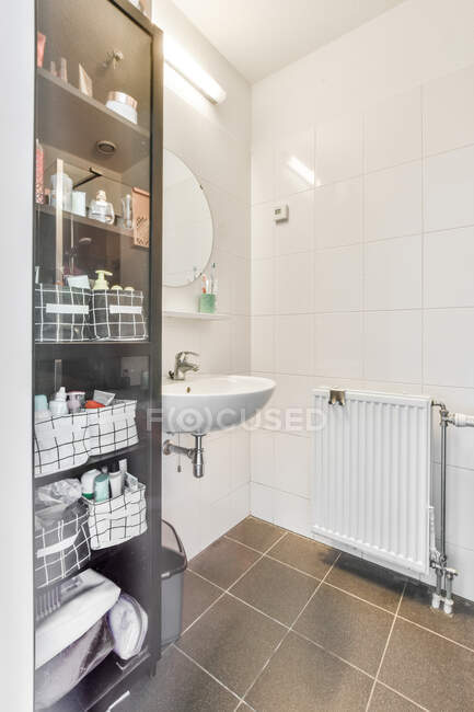 Assorted shower supplies placed in black cabinet with glass door near sink and mirror in light bathroom in apartment — Stock Photo