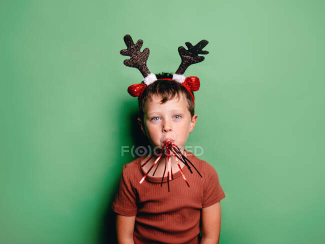 Niño usando cuernos de reno diadema y soplador fiesta festiva en la boca de pie sobre fondo verde y mirando a la cámara - foto de stock