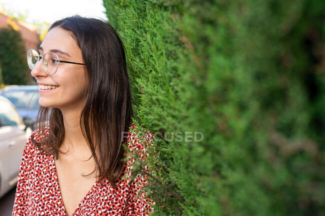 Cheerful young female with brown hair in eyeglasses standing among green branches and looking away in daylight — Stock Photo