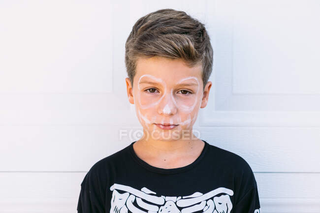 Serious preteen boy with white painted skeleton makeup dressed in black Halloween costume looking at camera against white wall — Stock Photo