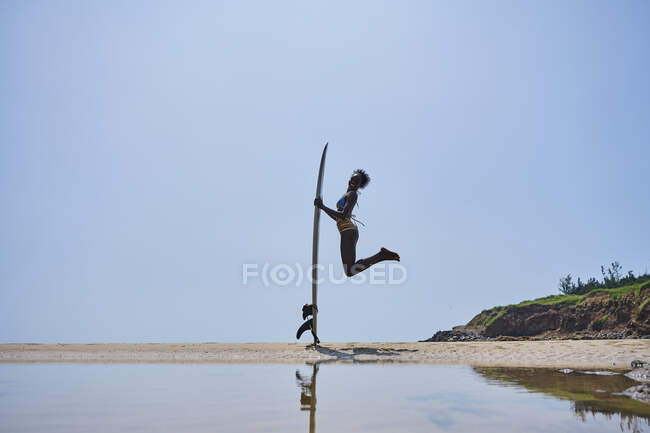 Side view of happy African American female surfer with longboard surf jumping above sandy coast under blue sky — Stock Photo