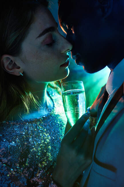 Вид збоку пари культур зі склом на шампанське в момент поцілунку проти блискучого світлого променя, дивлячись один на одного під час вечірки — стокове фото