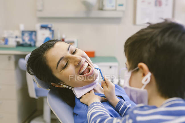 Menino curioso em máscara médica desempenhando papel de dentista e verificando dentes com espelho dentário no hospital — Fotografia de Stock