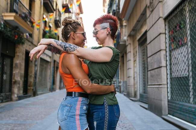 Вид збоку модна молода лесбійська пара з татуюваннями в сонцезахисних окулярах, що приховують погляд один на одного в момент поцілунку в місті — стокове фото