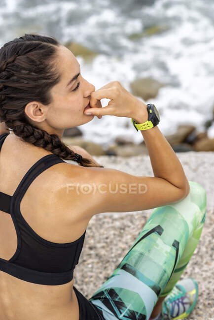 Vue latérale d'une athlète féminine mince en tenue de sport assise sur une promenade en pierre près de la mer et faisant une pause pendant l'entraînement — Photo de stock