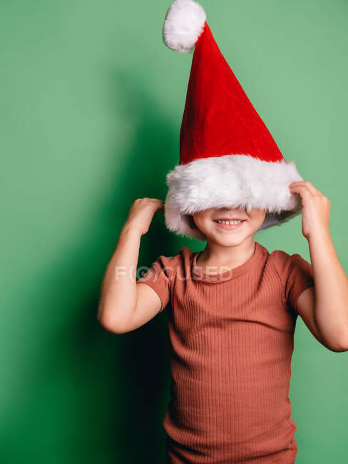 Неузнаваемый счастливый мальчик покрывает лицо красной шляпой Санты, стоящей на зеленом фоне. — стоковое фото