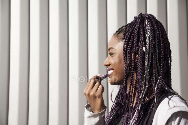 Side view of happy stylish young African American female with dyed purple Afro braids applying purple lipstick while standing against gray striped wall — Stock Photo