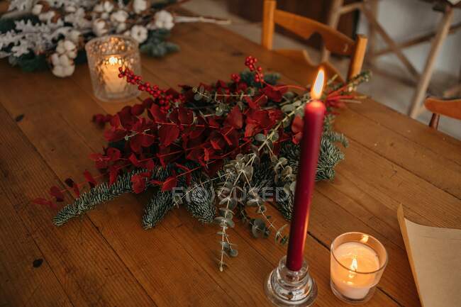 Сверху праздничный рождественский букет с ветвями хлопка, ели и веток эвкалипта и ярко-красными ветвями с ягодами, помещенными на деревянный стол со свечами в помещении — стоковое фото