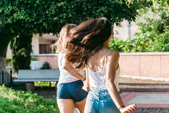 Vue arrière des meilleures amies anonymes se tenant la main tout en courant sur une allée carrelée en ville — Photo de stock