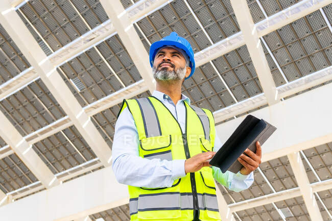 Low angle of middle aged Hispanic male engineer in uniform with tablet standing looking away under solar power station — Stock Photo