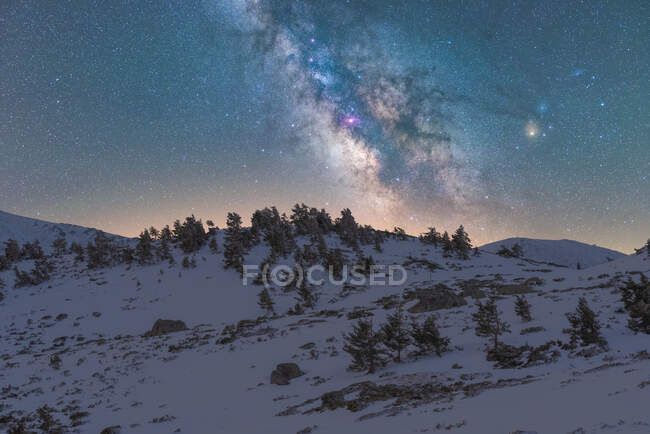 Atemberaubende Landschaft von schneebedeckten Hügeln und Bäumen vor hohen felsigen Bergen unter nächtlichem Sternenhimmel mit Milchstraße — Stockfoto