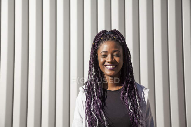 Feliz jovem afro-americano feminino com longas tranças tingidas sorrindo brilhantemente e olhando para a câmera enquanto se diverte contra a parede cinza na luz solar — Fotografia de Stock