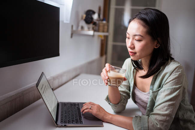 Side view of young ethnic female blogger at desk with netbook and coffee looking at camera in house room — Stock Photo
