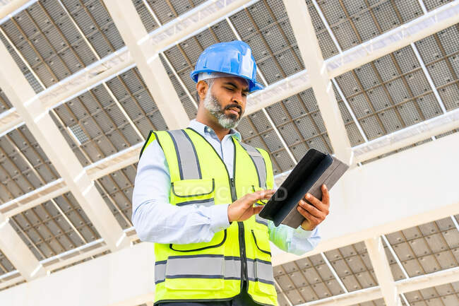 Angolo basso di mezza età ispanico ingegnere maschio in uniforme con tablet in piedi guardando schermo sotto la centrale solare — Foto stock