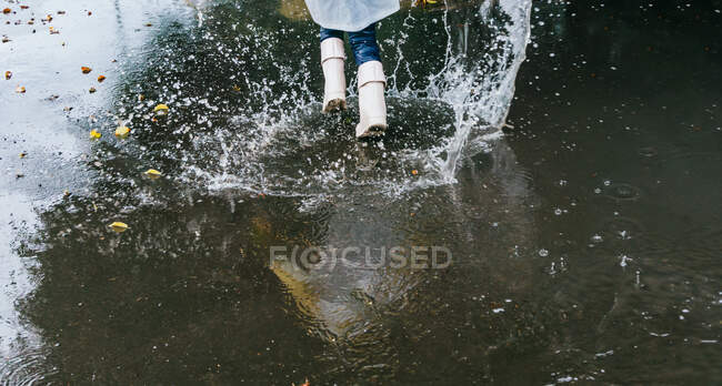 Desde arriba vista trasera de la cosecha niño anónimo en botas de goma que se divierten en charco con salpicaduras de agua en el día lluvioso - foto de stock