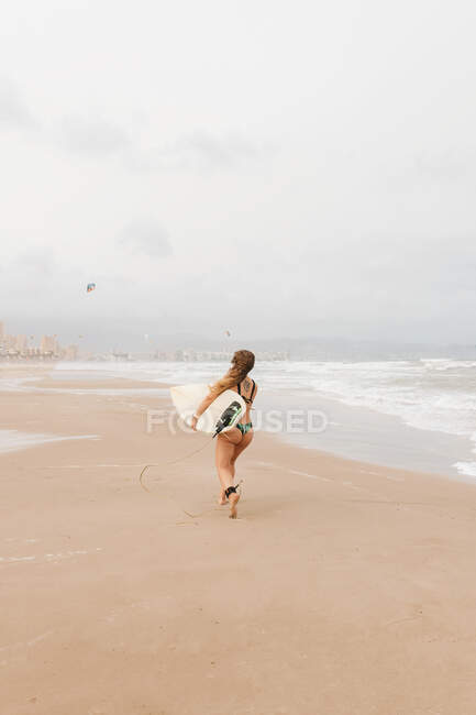 Back view of unrecognizable young sportswoman in swimwear with surfboard looking away on sandy coast against stormy ocean — Stock Photo