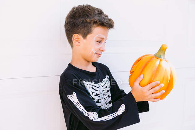 Allegro ragazzo preadolescente in costume scheletro con trucco con zucca arancione intagliata mentre si prepara per la festa di Halloween contro il muro bianco — Foto stock