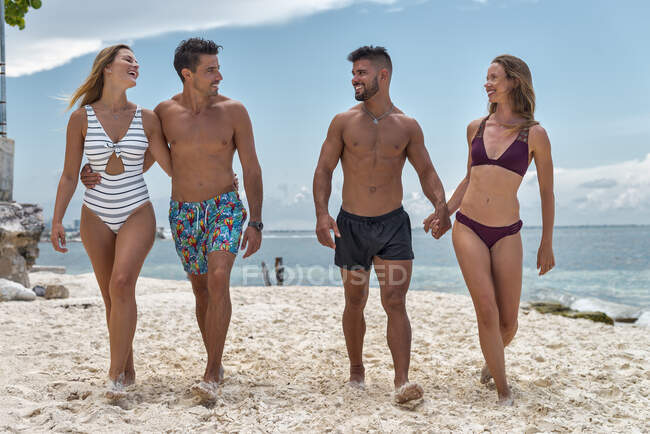Cheerful tourists in swimwear talking while strolling on sandy shore against ocean under cloudy blue sky in town — Stock Photo