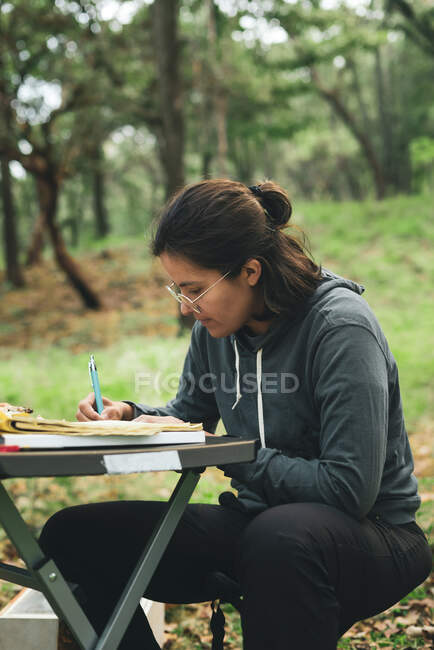 Концентрована жінка сидить за столом і робить нотатки в блокноті в зеленому парку вдень — стокове фото