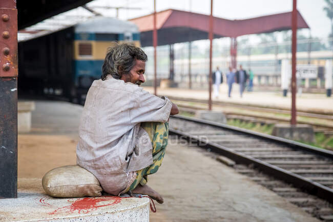 INDIA, BANGLADESH - DECEMBER 6, 2015: Side view of ethnic male in worn out dirty clothes sitting on crouched on platform of railway station and looking away — Stock Photo