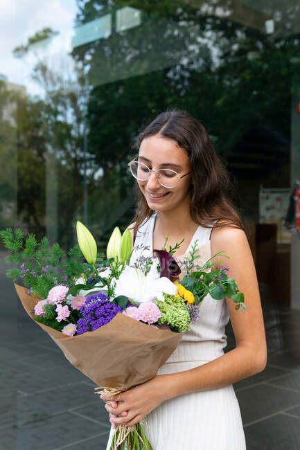 Charming young female in eyeglasses with blooming flower bouquet against glass wall in town in daylight — Stock Photo