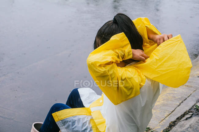 Vista lateral de criança anônima colocando no capuz de slicker enquanto sentado na passarela em tempo chuvoso — Fotografia de Stock