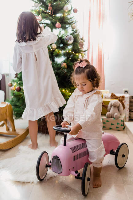 Full body of little cute girl riding velomobile near sister decorating Christmas tree with baubles during holiday celebration — Stock Photo