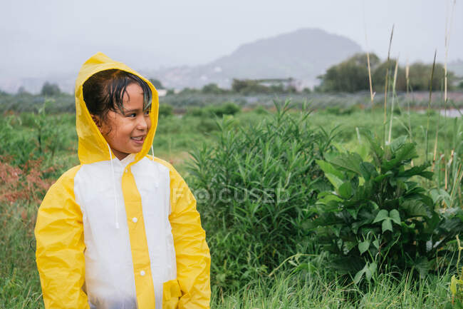 Charming ethnic child in slicker looking away against tropical plants on meadow in rainy weather — Stock Photo