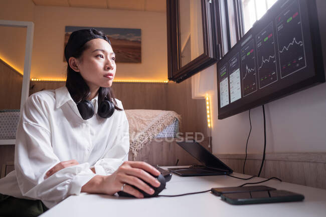 Vista lateral de mujeres asiáticas concentradas trabajando en computadora con gráficos que muestran la dinámica de los cambios en el valor de la criptomoneda en el lugar de trabajo conveniente - foto de stock