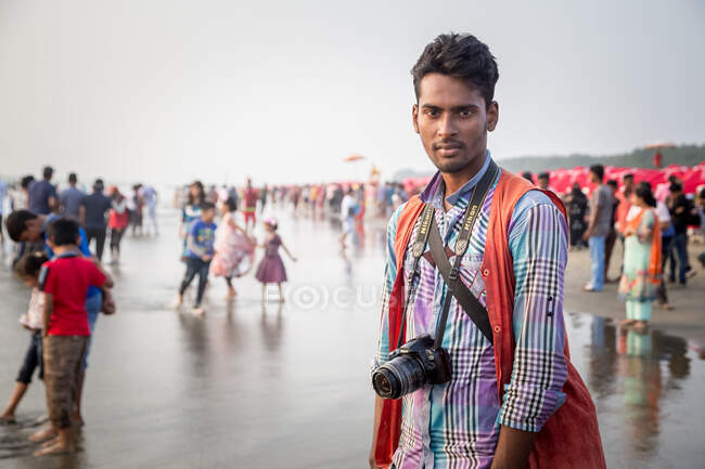 INDIA, BANGLADESH - 4 DE DICIEMBRE DE 2015: Joven hombre étnico con ropa casual de pie con cámara de fotografía profesional en arena mojada en la playa mirando a la cámara - foto de stock