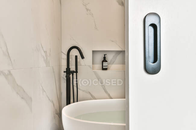 Porte ouverte à la salle de bain lumineuse avec murs en marbre et accessoires de salle de bain noir au-dessus de la baignoire remplie d'eau — Photo de stock
