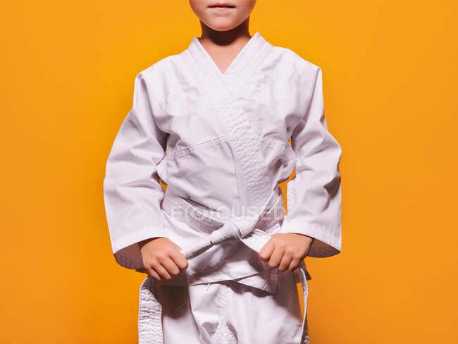 Niño serio anónimo en kimono blanco apretando nudo de cinturón de karate con sus manos sobre fondo naranja brillante y mirando a la cámara - foto de stock