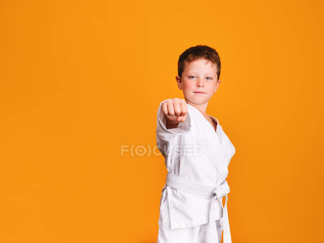 Self assured boy in white kimono doing karate punch and looking at camera with smile gaze against orange background — Stock Photo