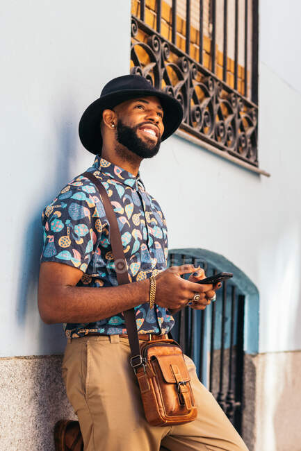 Vista lateral de la sonriente moda étnica masculina en el sombrero con el bolso de hombro de mensajes de texto en el teléfono celular en la ciudad soleada - foto de stock