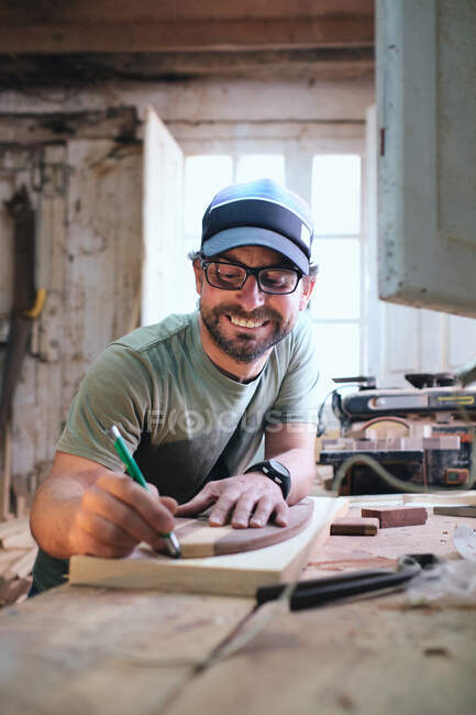 Obrero barbudo alegre en gafas y forma de patineta de seguimiento de gorra con marcador en tablón de madera en el taller - foto de stock