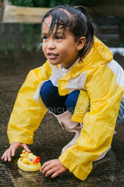 Charming ethnic kid in raincoat playing with plastic ducks reflecting in rippled puddle in rainy weather — Stock Photo